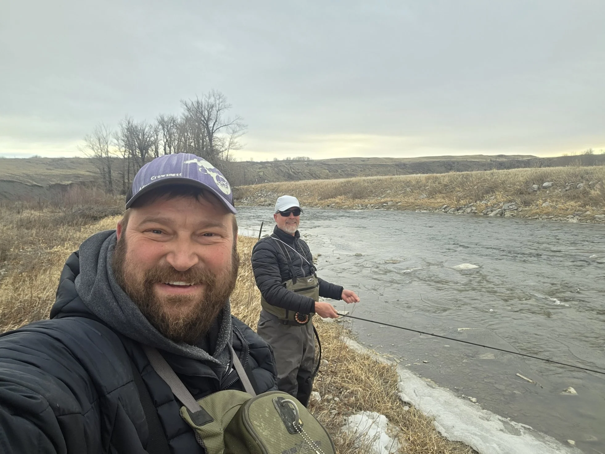Two men fishing by a riverbank in a rural area with leafless trees and hills in the background on a cloudy day.