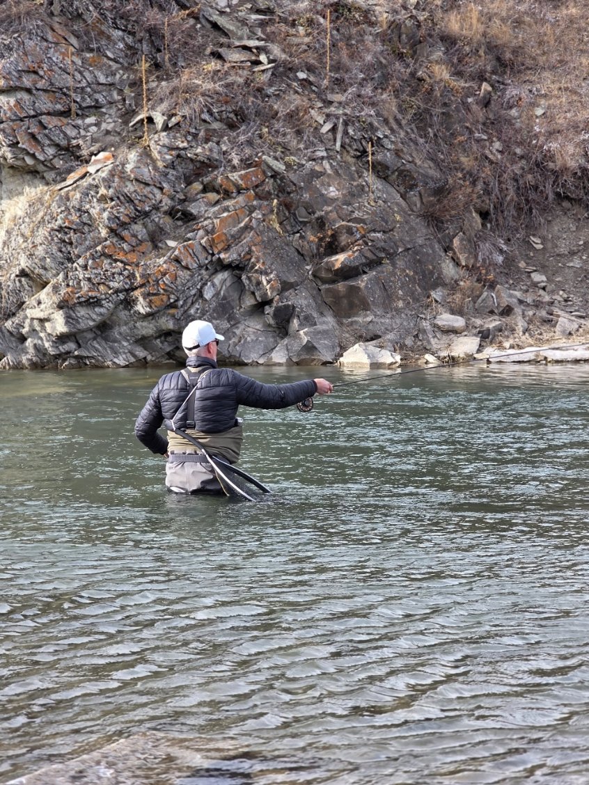A man standing in a river, fishing with a rod, wearing a black jacket, light-colored waders, and a white cap, with rocky cliffs and dry vegetation in the background.