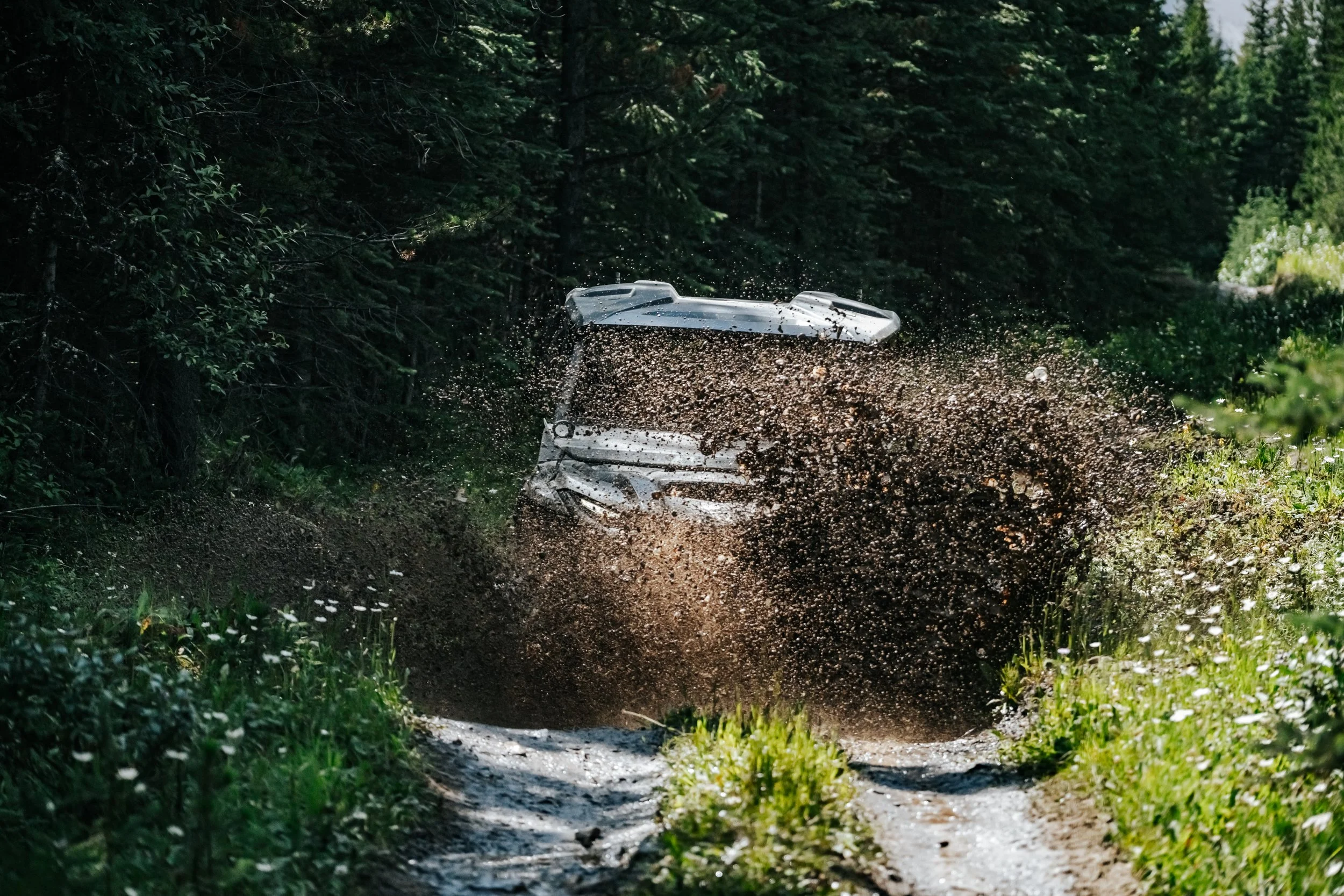 An off-road vehicle (Yamaha Rmax 4) splashing through mud on a forest trail with green trees and bushes surrounding the path.