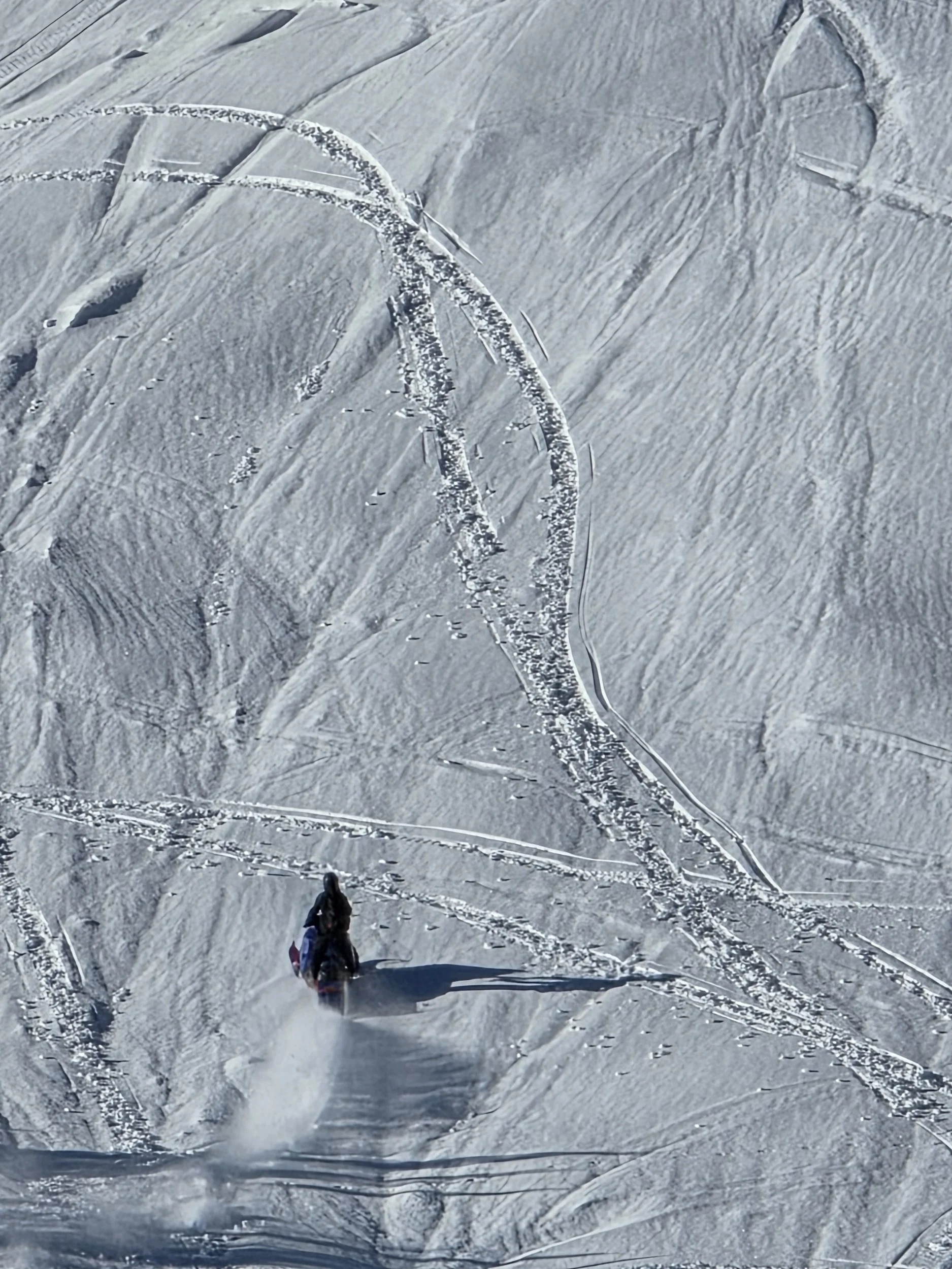 A snow-covered mountain landscape with ski tracks and two climbers at the bottom. The snow appears icy and textured.