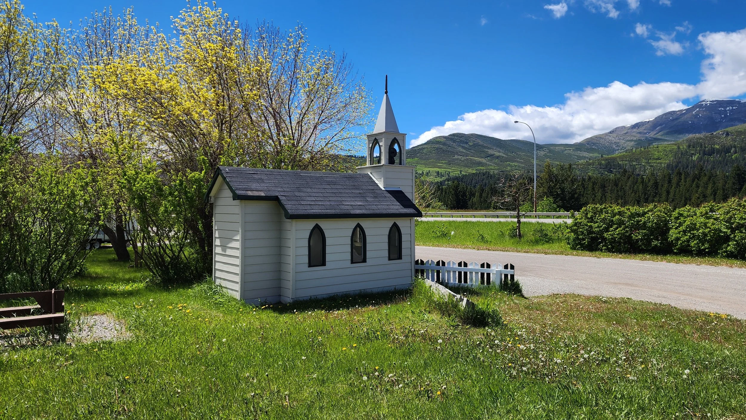 A small white church with a black roof and steeple, situated on a grassy area with trees and mountains in the background, under a partly cloudy blue sky.