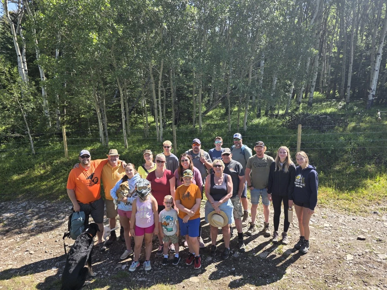 Group of people hiking in a forest with trees and greenery, some wearing outdoor gear and sunglasses, accompanied by a dog.