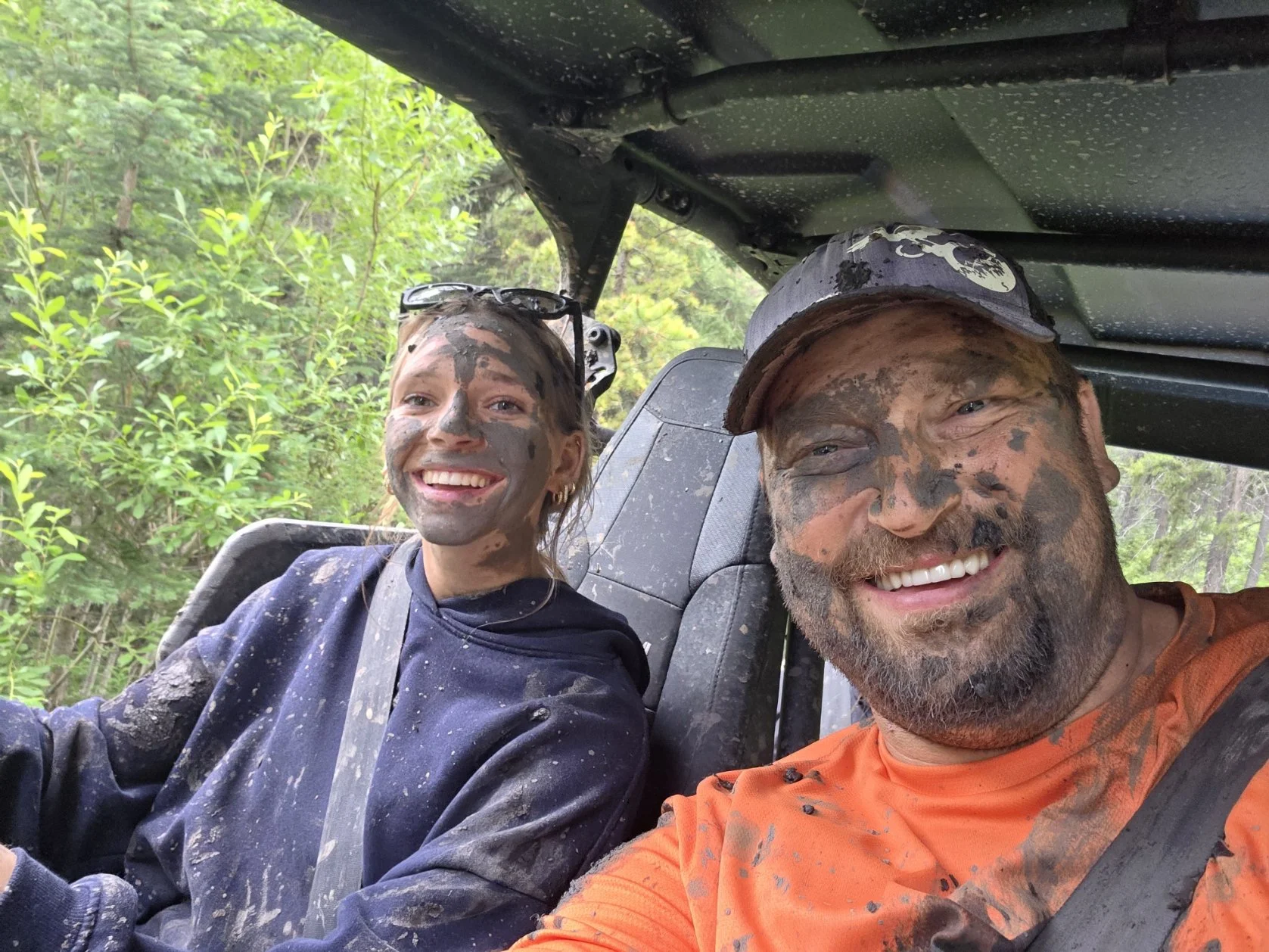 Two people smiling inside a vehicle, covered in mud, with a background of green trees and foliage.