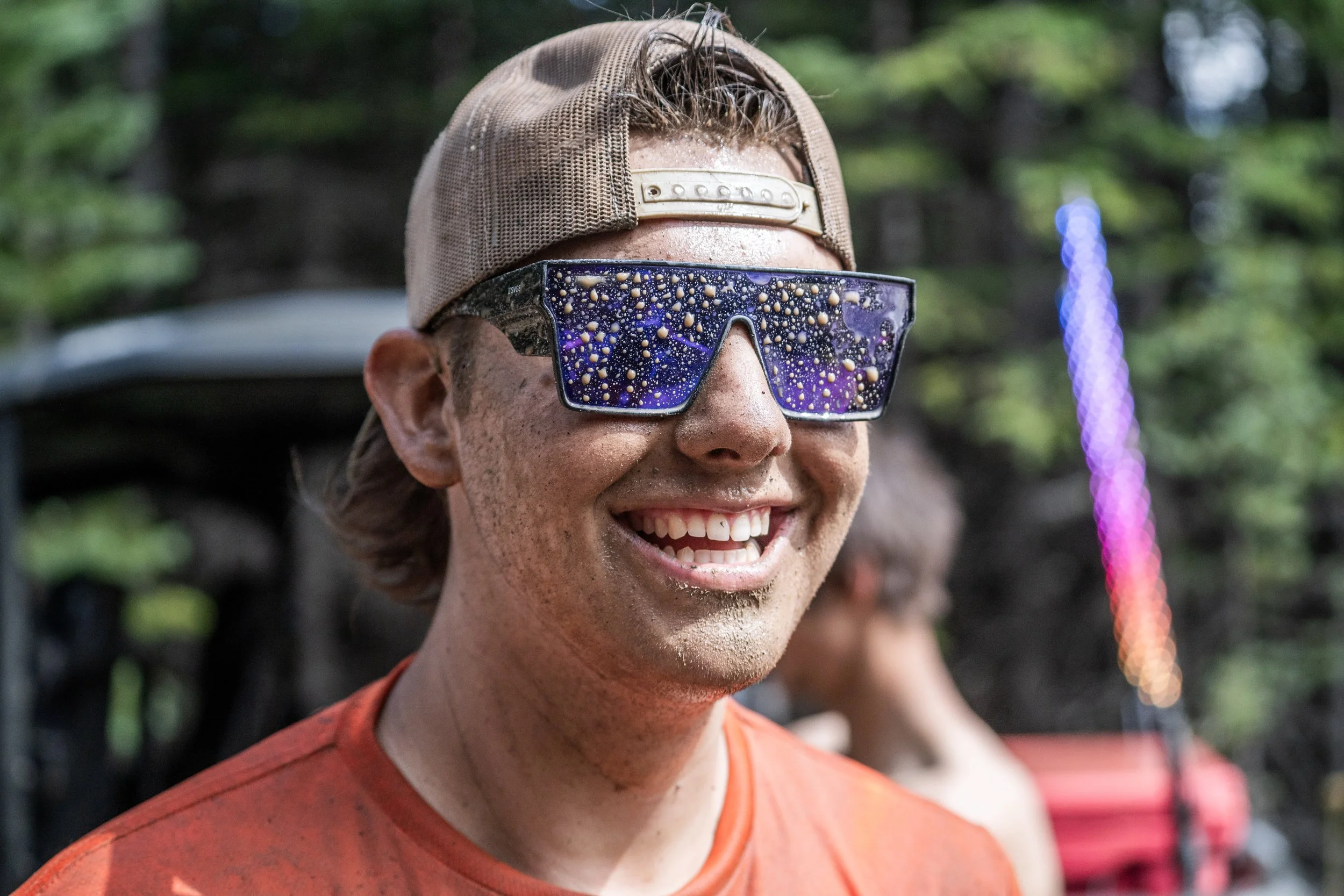 A smiling man wearing a khaki cap and futuristic sunglasses with a starry night pattern reflected on the lenses. He is outdoors, in a setting with trees and colorful lights in the background.