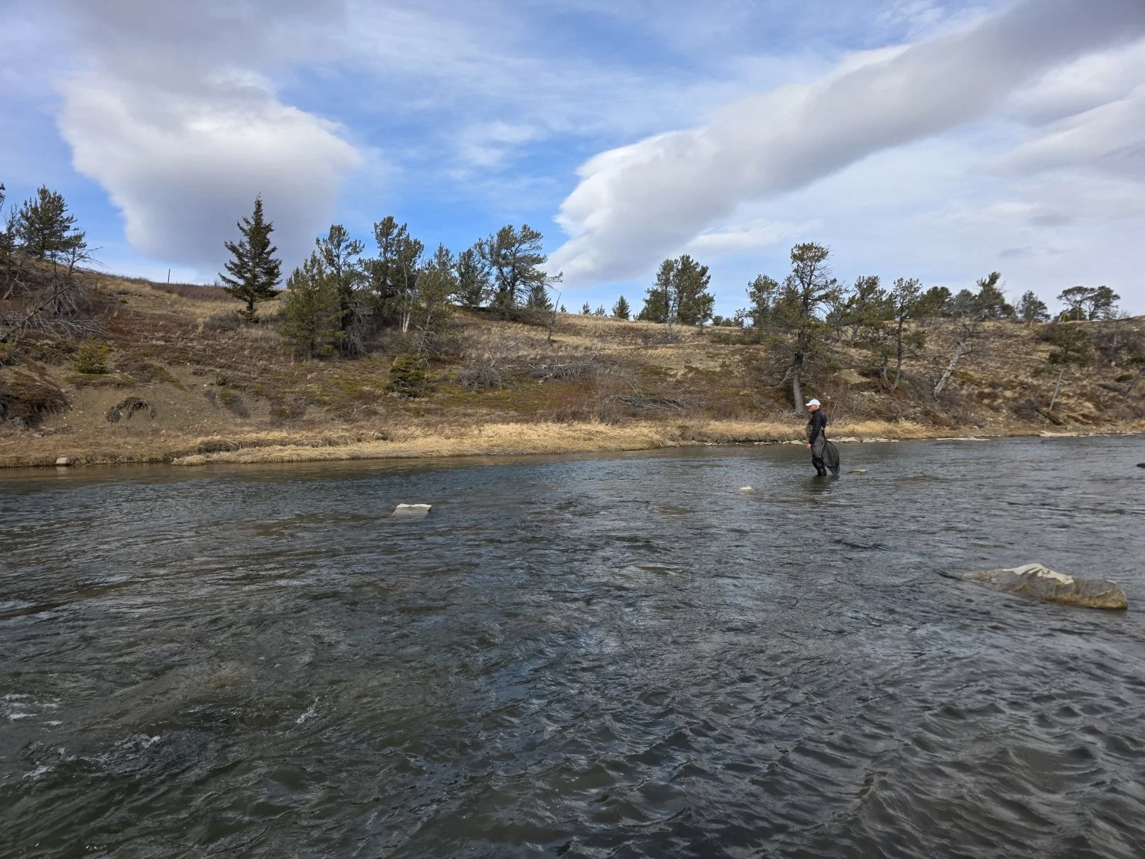 A person standing in a river with trees and hillside in the background under a partly cloudy sky.