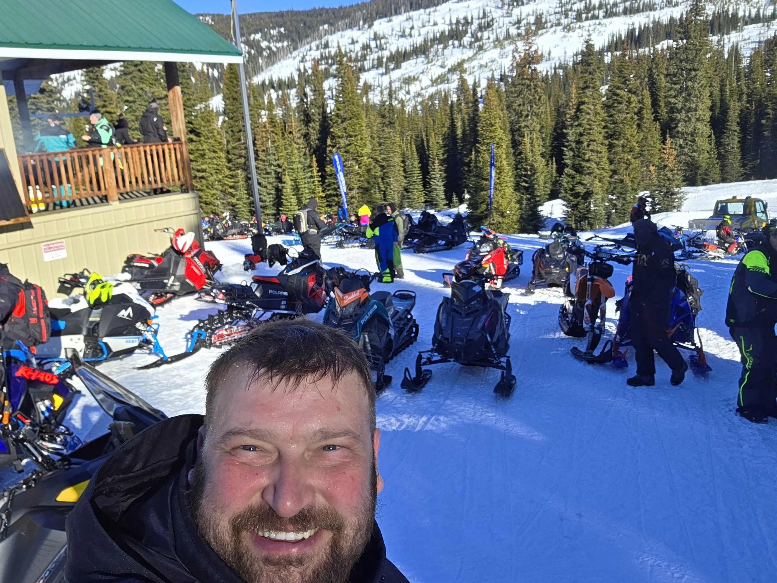 A man with a beard smiling at the camera in a snowy mountain area with parked snowmobiles, people in winter gear, and a green-roofed building. Pine trees and snow-covered slopes are in the background.