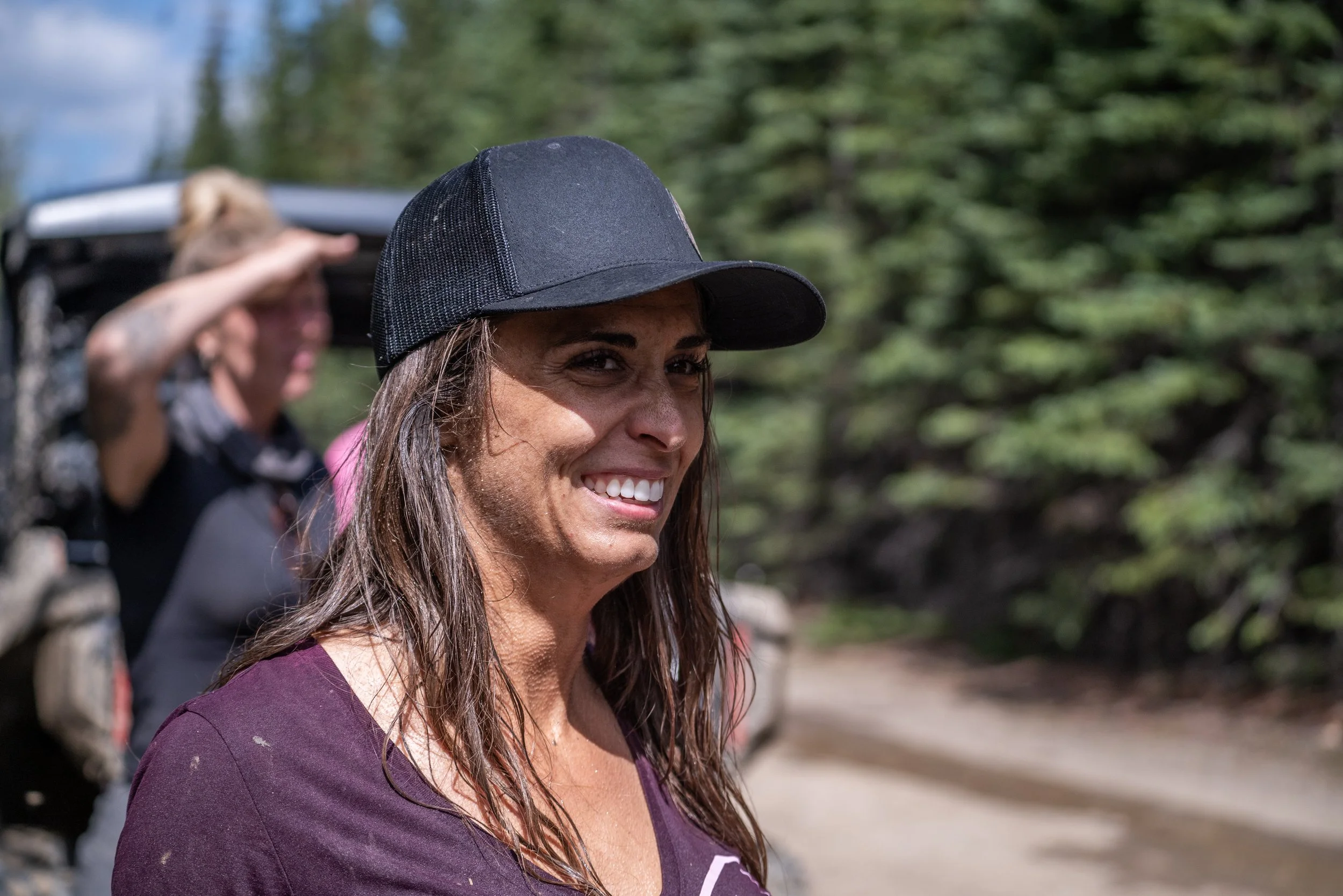 A woman with long brown hair wearing a black baseball cap, smiling outdoors with trees and a dirt road in the background.