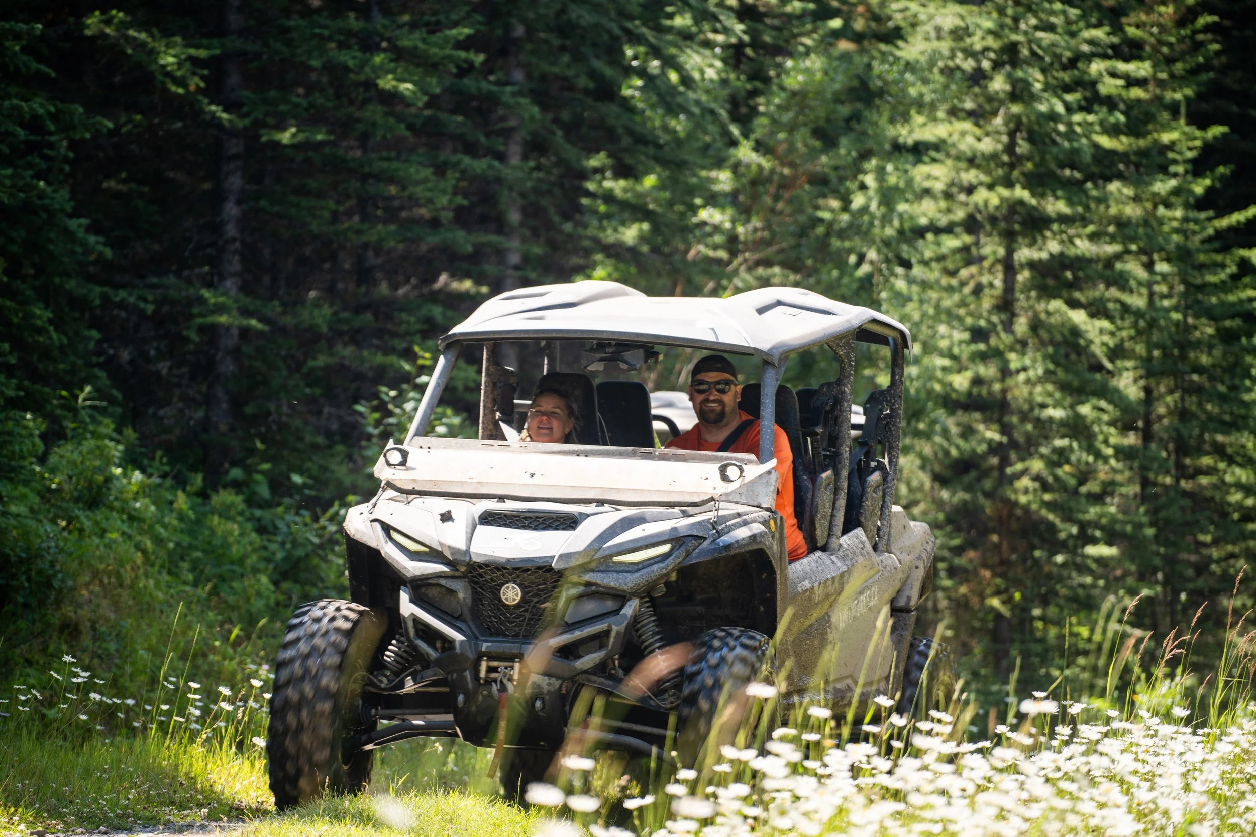 Two people riding an off-road utility vehicle (Yamaha Rmax 4) through a lush, green forested area on a sunny day.
