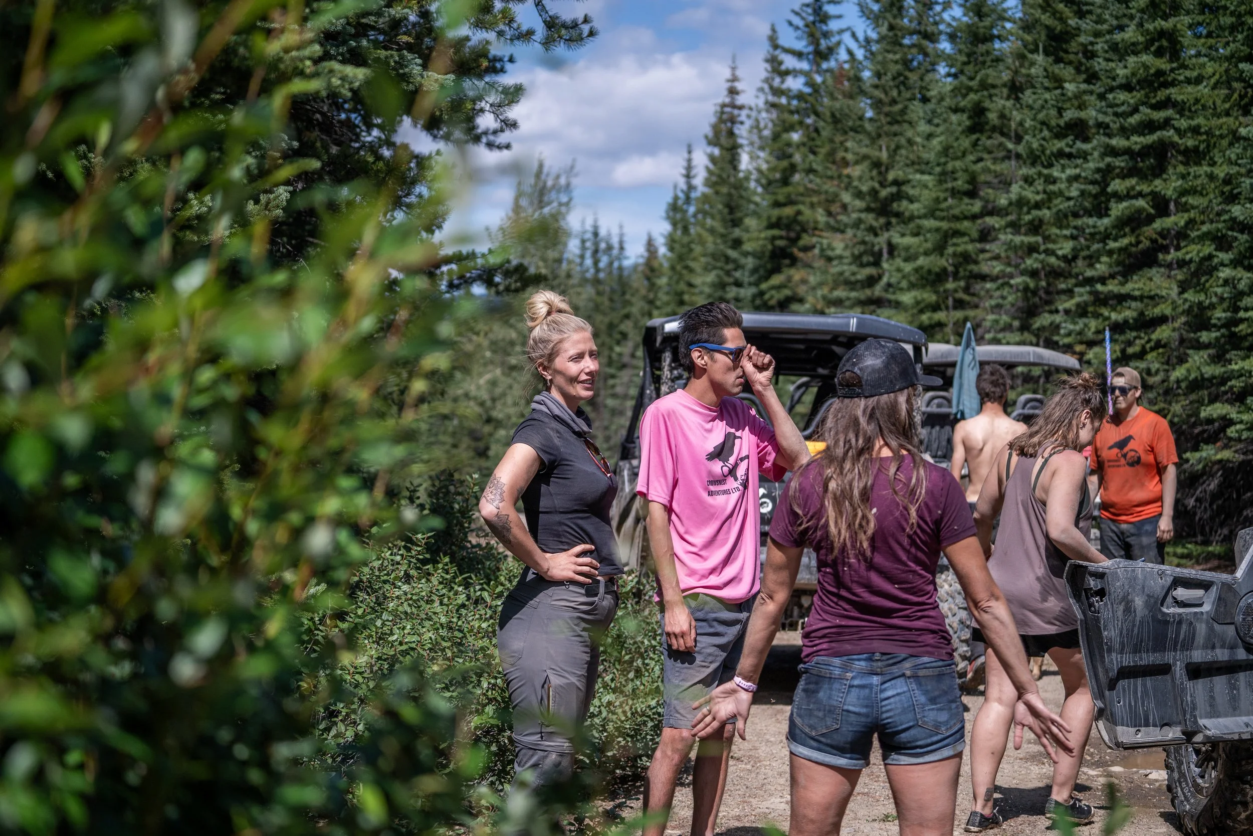 Group of people outdoor in a wooded area, some near a golf cart, engaged in conversation or preparing for activities, with trees and clouds in the background.