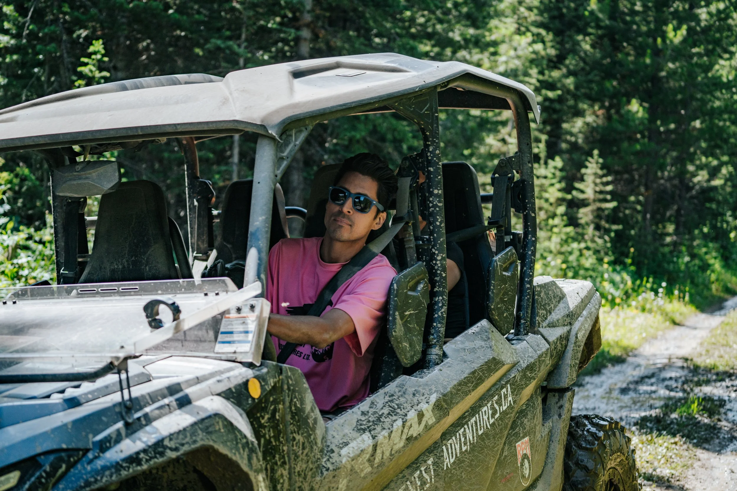 A man wearing sunglasses and a pink T-shirt sitting in a muddy off-road utility vehicle on a dirt trail surrounded by green trees.