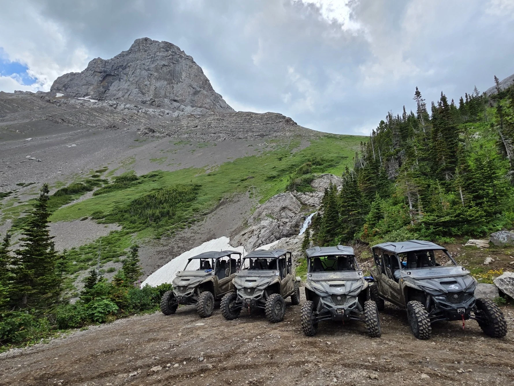 Four all-terrain utility vehicles parked on a dirt trail in a mountainous area with green trees, a rocky slope, and a waterfall in the background.