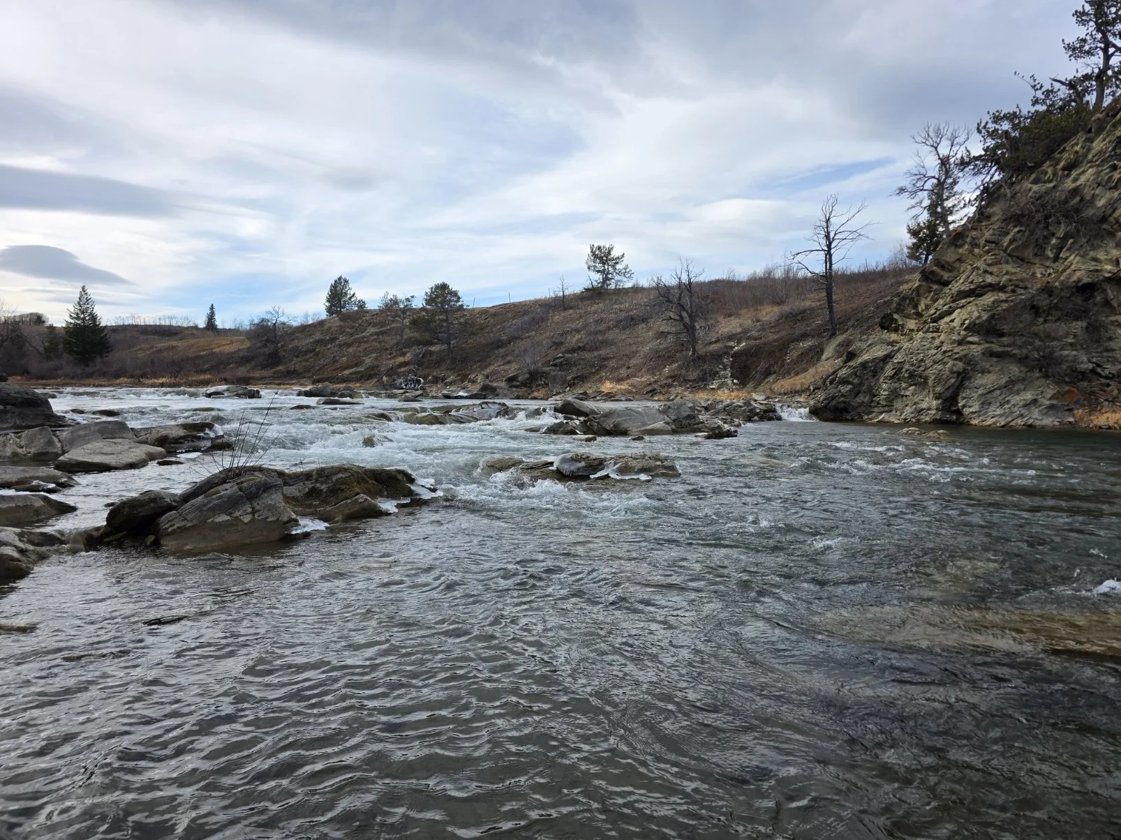 A flowing river with rocks along the banks, surrounded by hills and sparse trees, under a cloudy sky.
