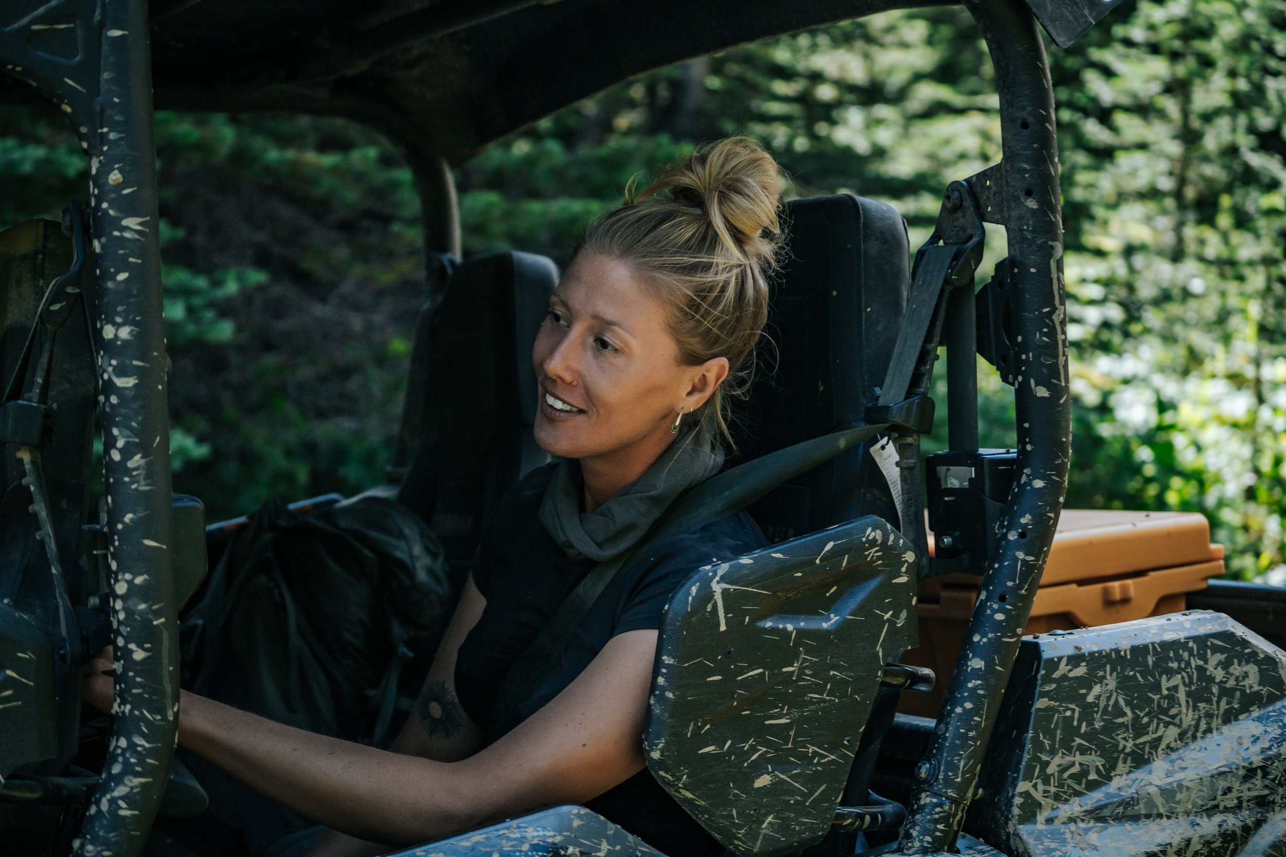 A woman with her hair in a bun sitting in a muddy, off-road vehicle in a wooded area, looking to her right and smiling.