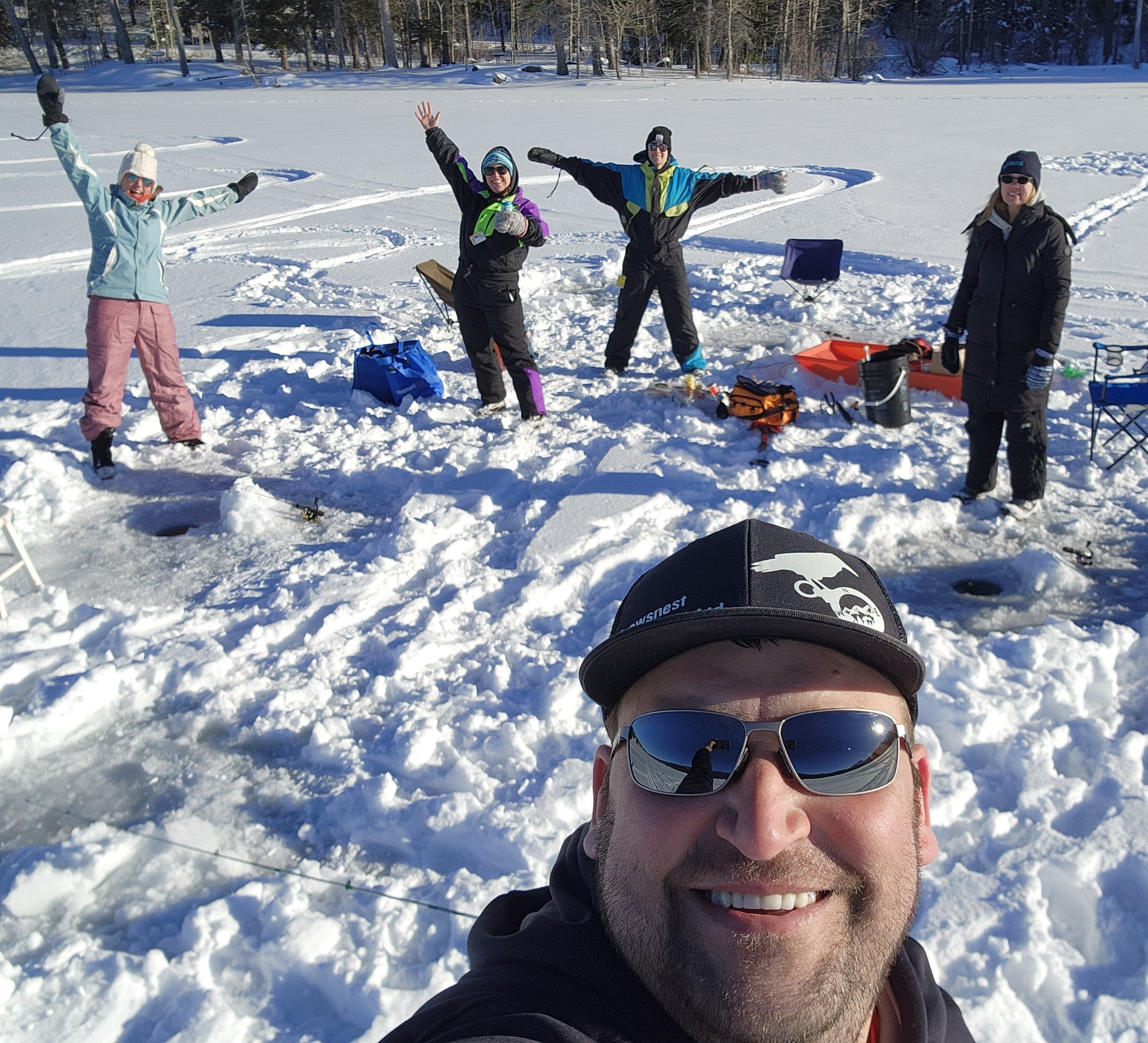 Group of people on a snowy lake, some with fishing gear, wearing winter clothes, posing for a selfie, with a man in the foreground wearing sunglasses and a black cap, reflecting the snow and sky.