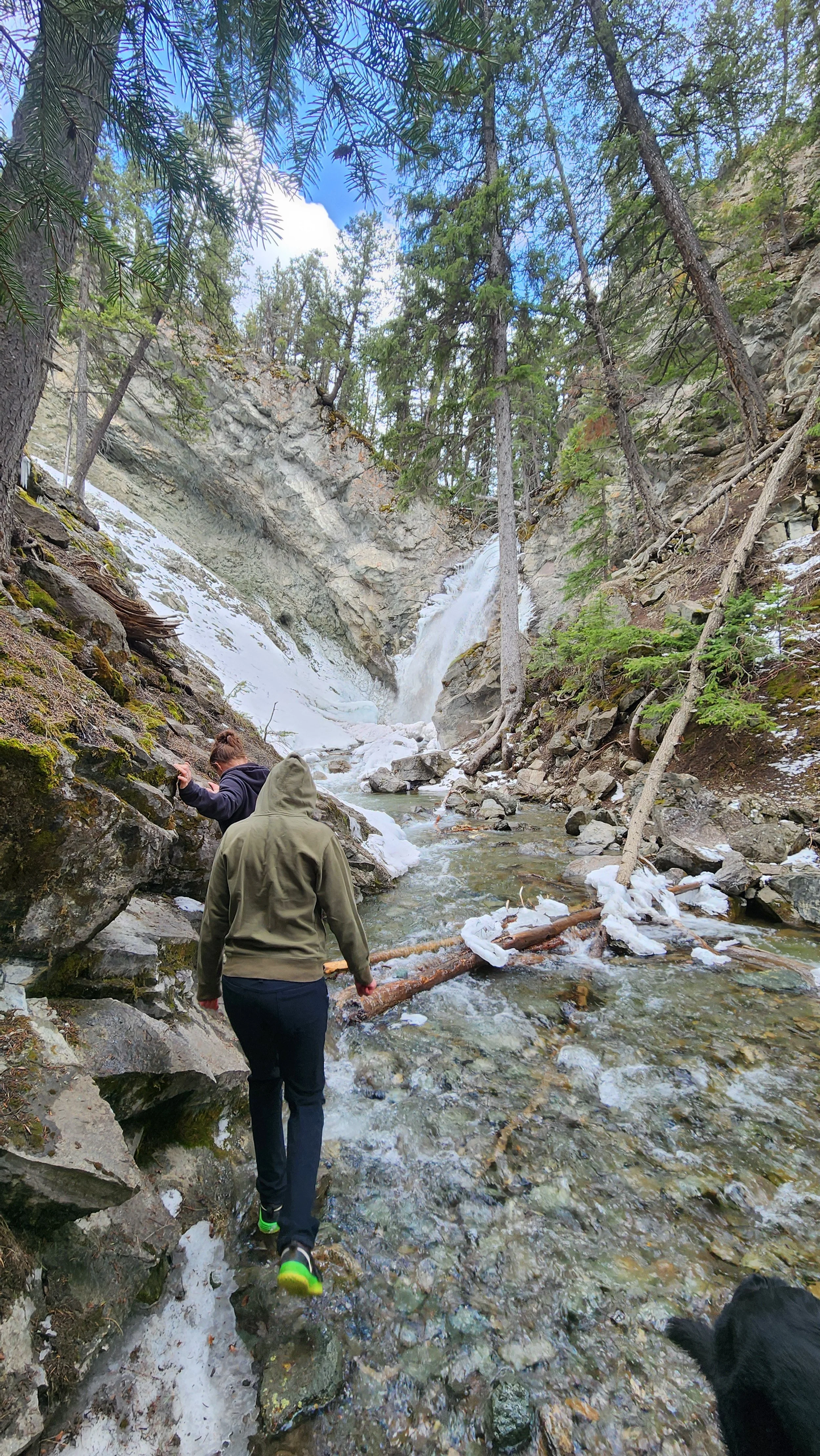 Two people hiking in a forest near a waterfall, with patches of snow and ice on the ground and rocks.