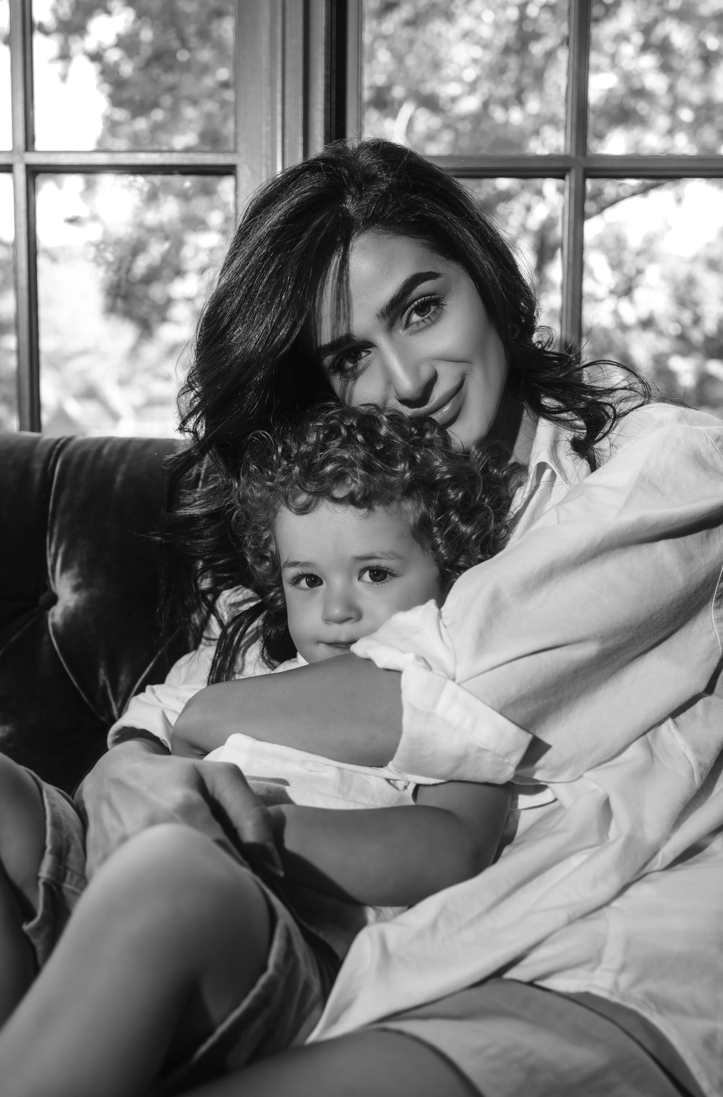 A woman and a young boy with curly hair sitting close together on a couch, smiling softly. The woman has dark hair, and the boy has a serious expression. They are in a room with large windows in the background.
