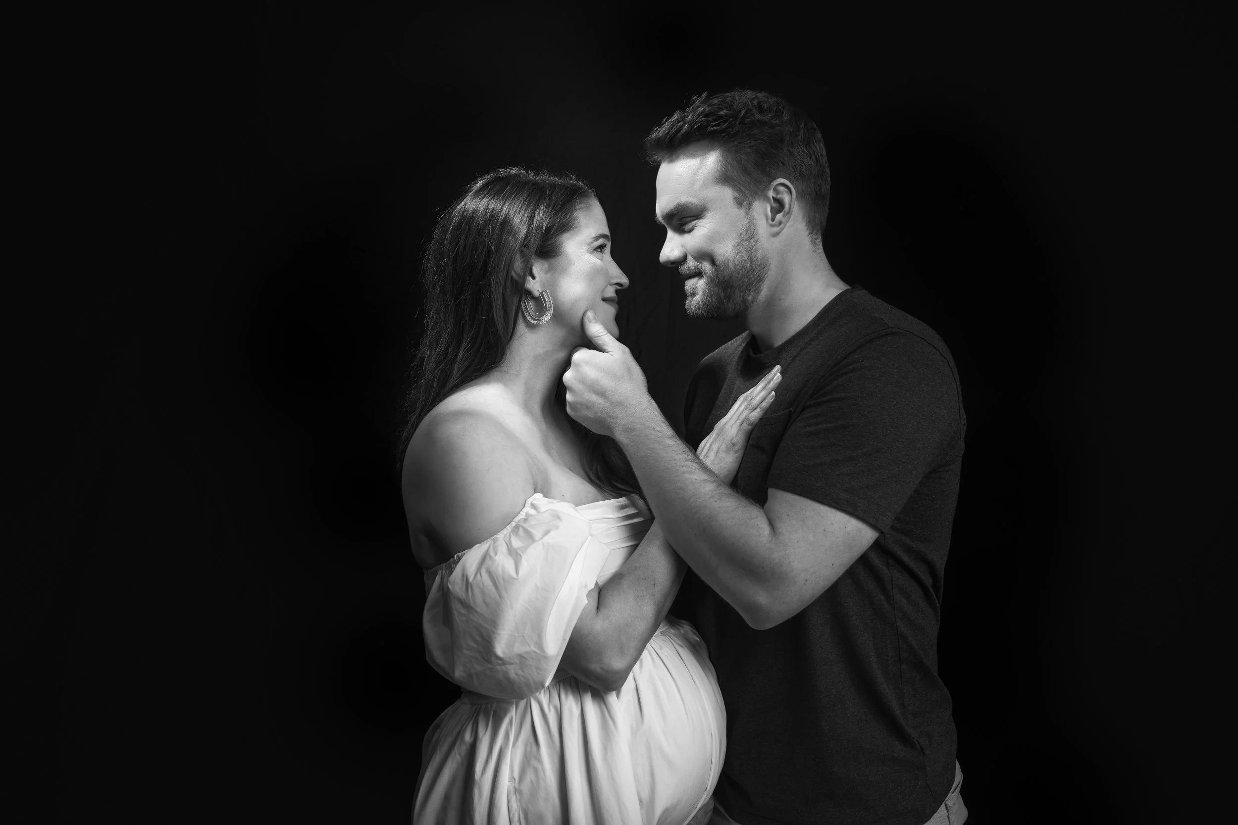 A man and a pregnant woman looking at each other affectionately, with the man's hand gently holding her chin and her hand resting on his chest, against a black background.