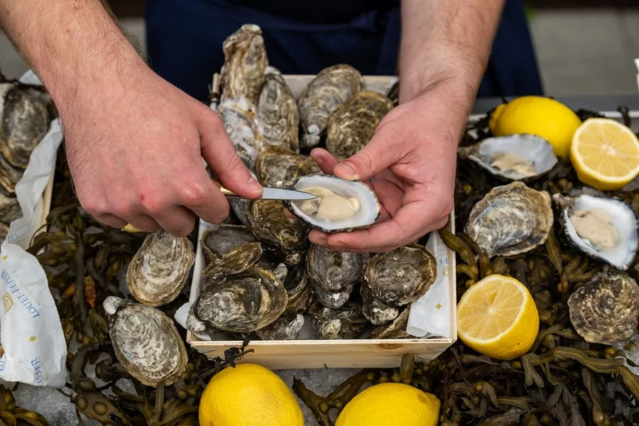 Person shucking oysters on a tray surrounded by lemons and seaweed.