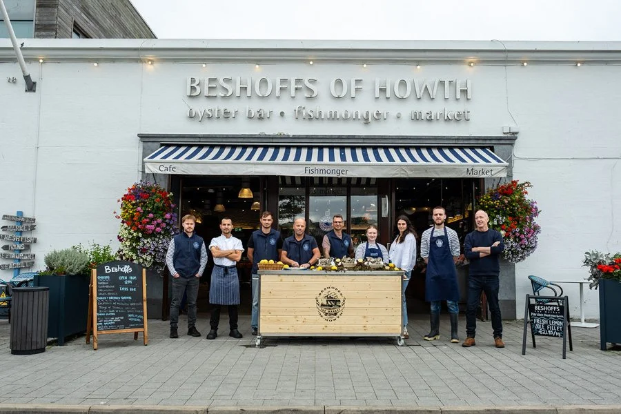 Group of nine people standing in front of a seafood market and cafe named 'Beshofs of Howth', with a seafood display table and planters with colorful flowers on either side.