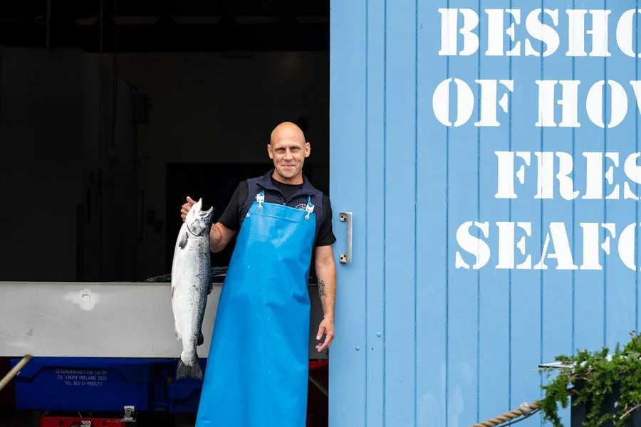 A man in a blue apron holding a large fish, standing outside a seafood store.