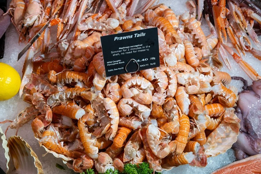 Fresh peeled prawn tails on ice at a seafood market, with a sign indicating they are Atlantic Nephrops norvegicus, also known as Norway lobster, with prices listed.