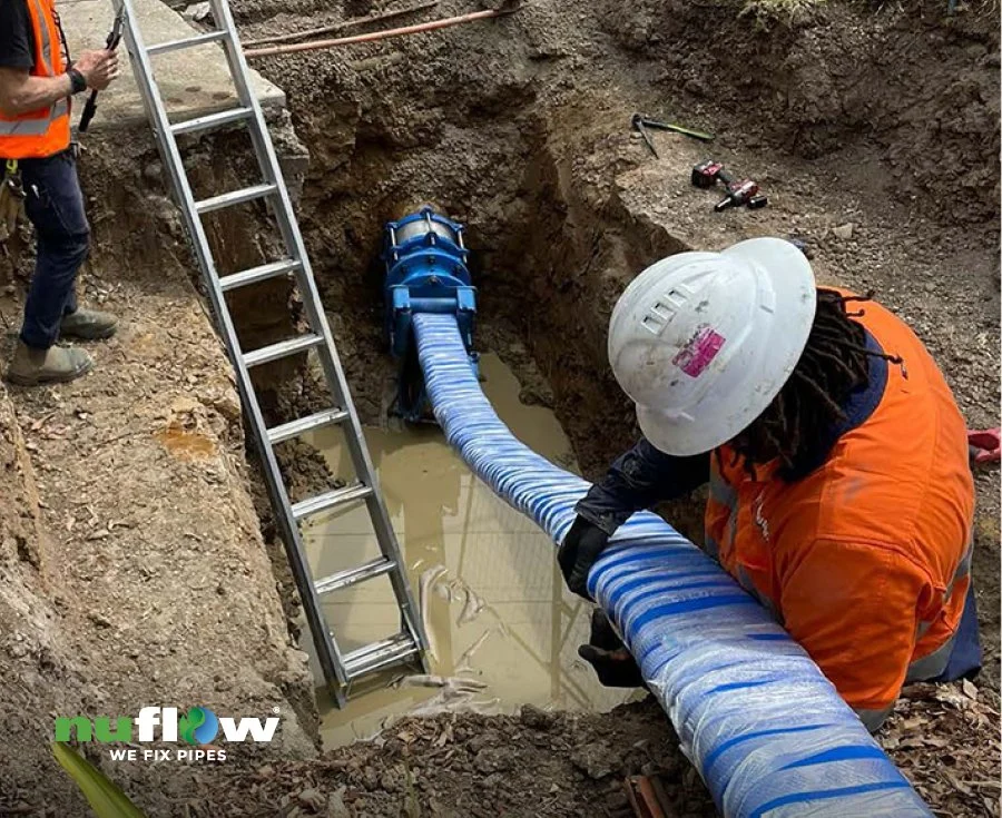 Workers installing or repairing a pipeline in a trench, using a blue pipe liner. One worker is holding the pipe, wearing an orange vest and a hard hat. A ladder is positioned nearby for access.