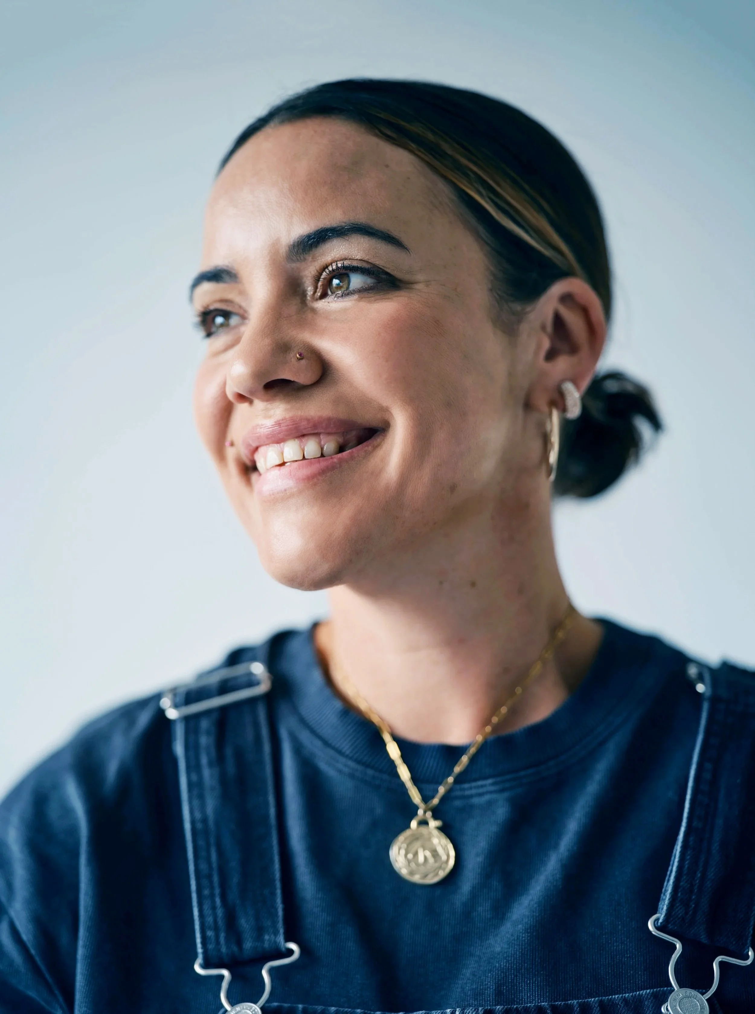 A smiling woman with brown eyes and dark hair tied back, wearing a navy blue top or dress, gold earrings, a gold chain necklace with a coin pendant, and a nose piercing, looking slightly to her left.