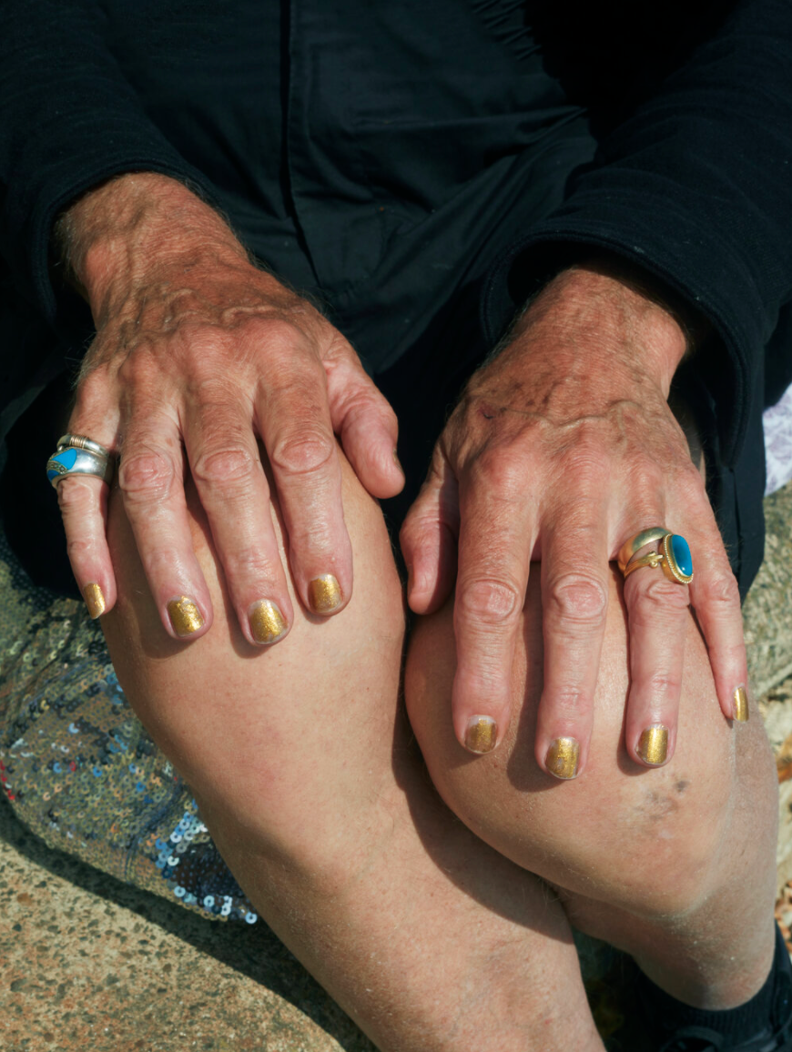Close-up of a person's hands resting on their knees, showing gold painted nails and multiple rings, with a black long-sleeve shirt. The person is sitting on the ground with a textured surface beneath.
