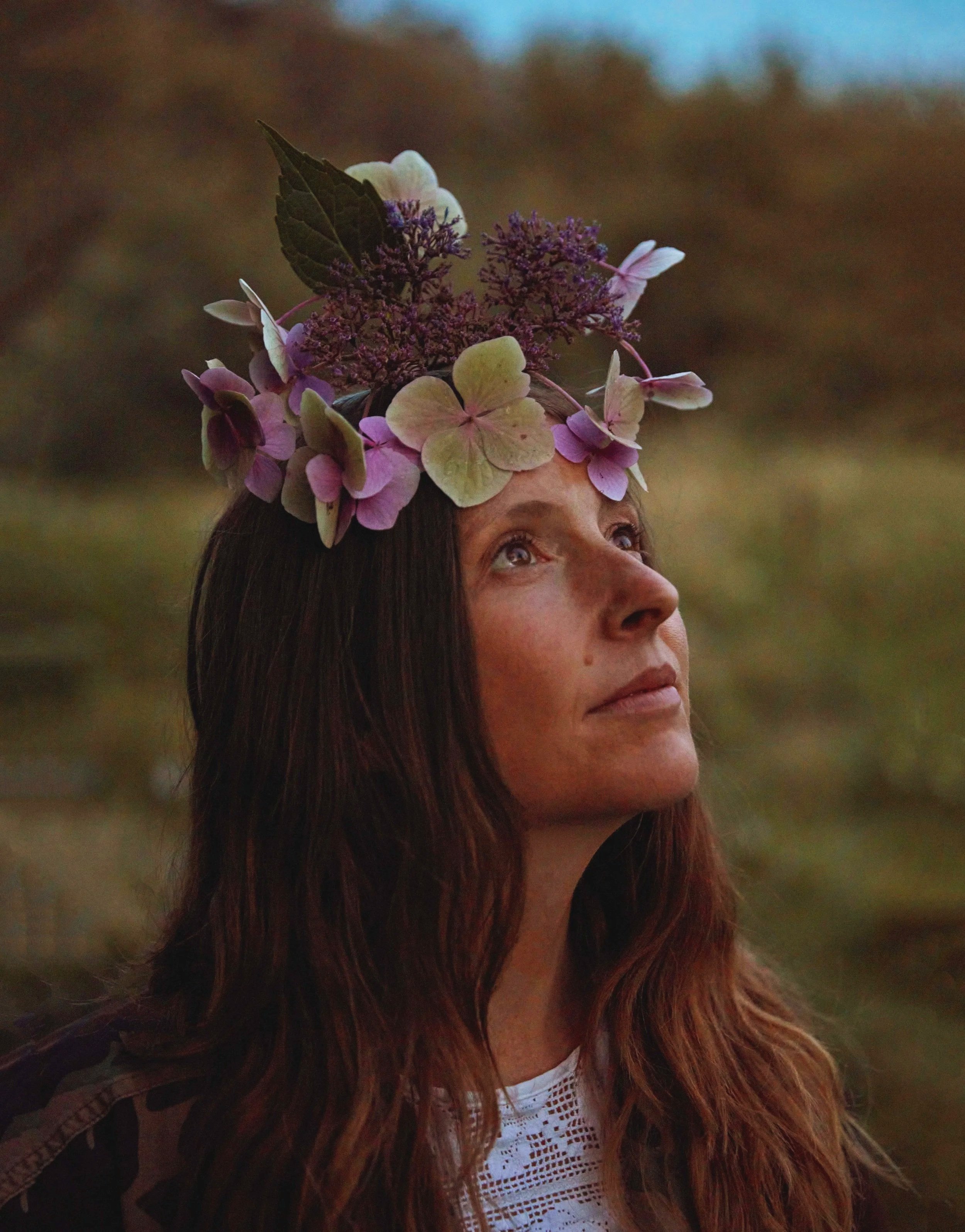 A woman with long brown hair wearing a floral crown made of pink, purple, and white flowers, looking upwards outdoors during dusk.