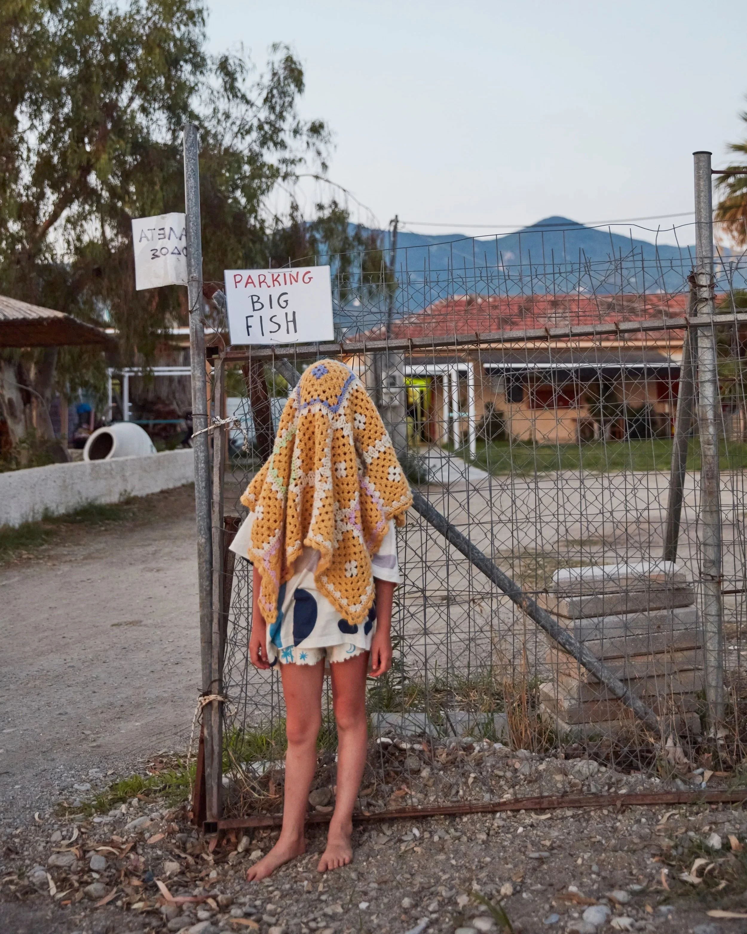 Person standing barefoot in front of a wire gate with signs, mountains in the distance, and a blanket draped over the person's shoulders.