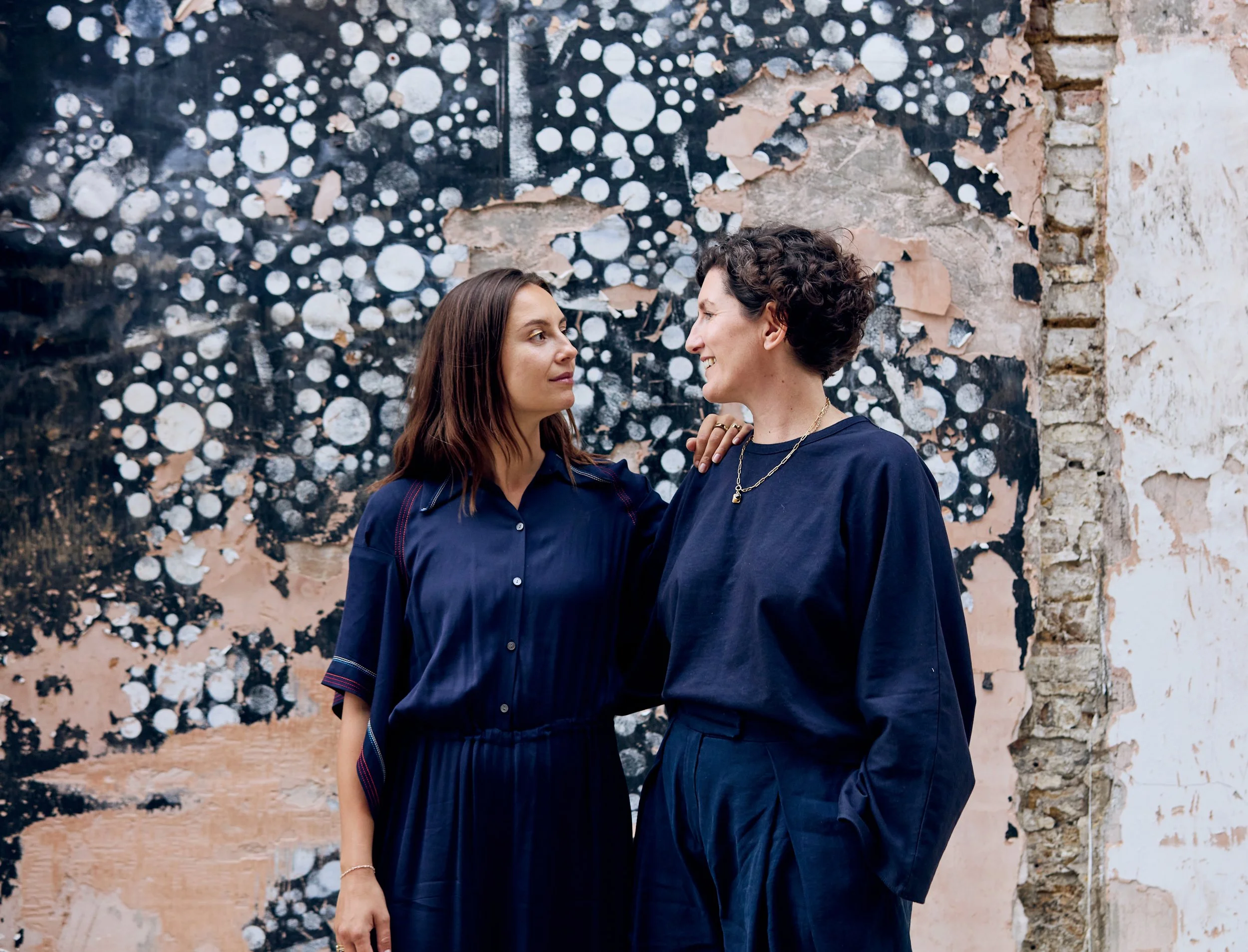Two women standing close to each other and looking into each other's eyes against a textured brick wall with black and white circular artwork.