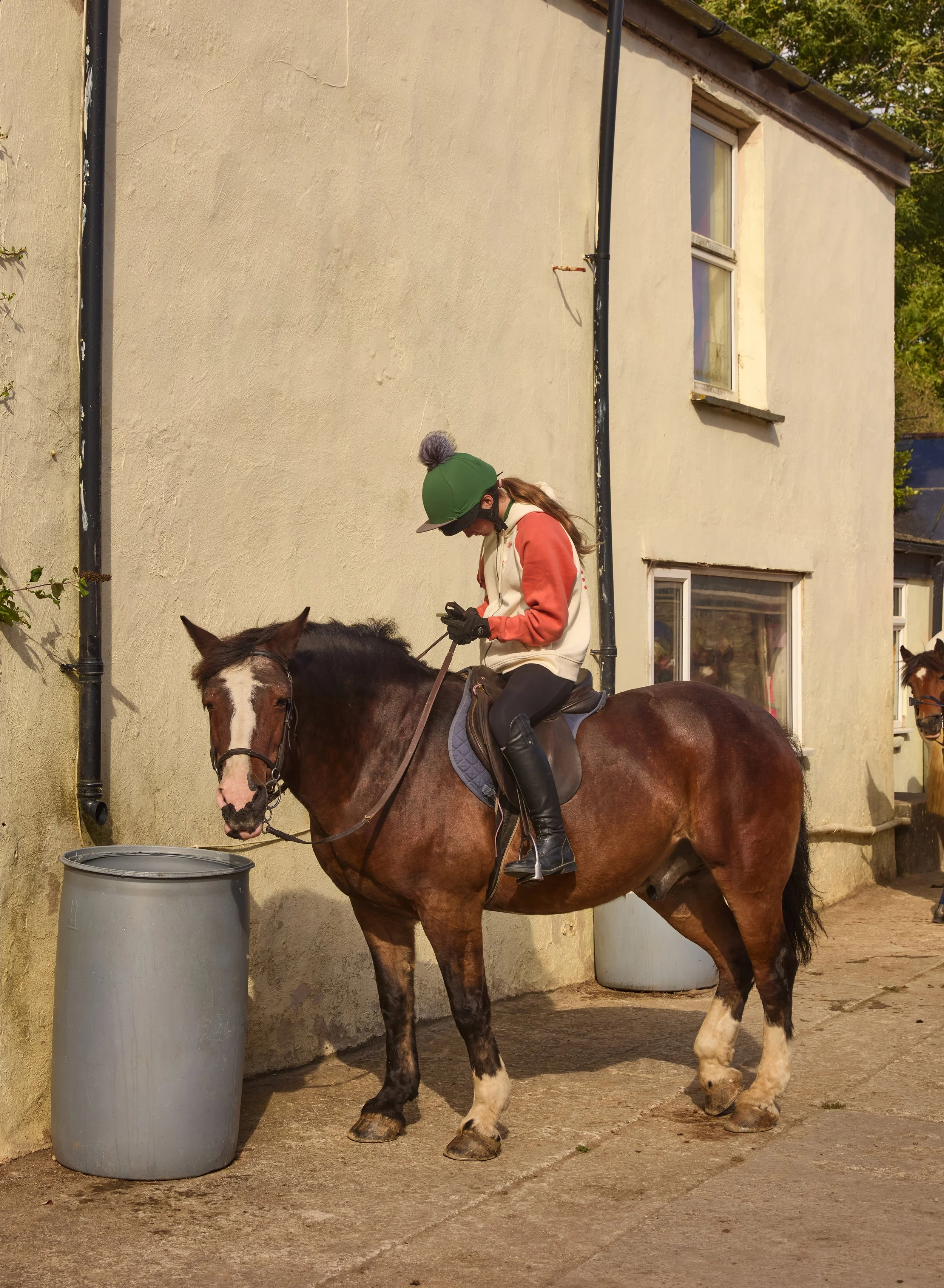 A person on horseback at a stable, wearing a green helmet, pink and white sweatshirt, black gloves, and sunglasses, looking at their phone, with a building wall behind them.