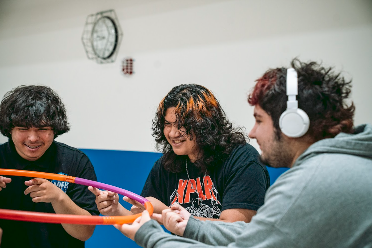 Three young adults playing with colorful pool noodles, smiling and wearing casual clothes, in an indoor room with a wall clock.
