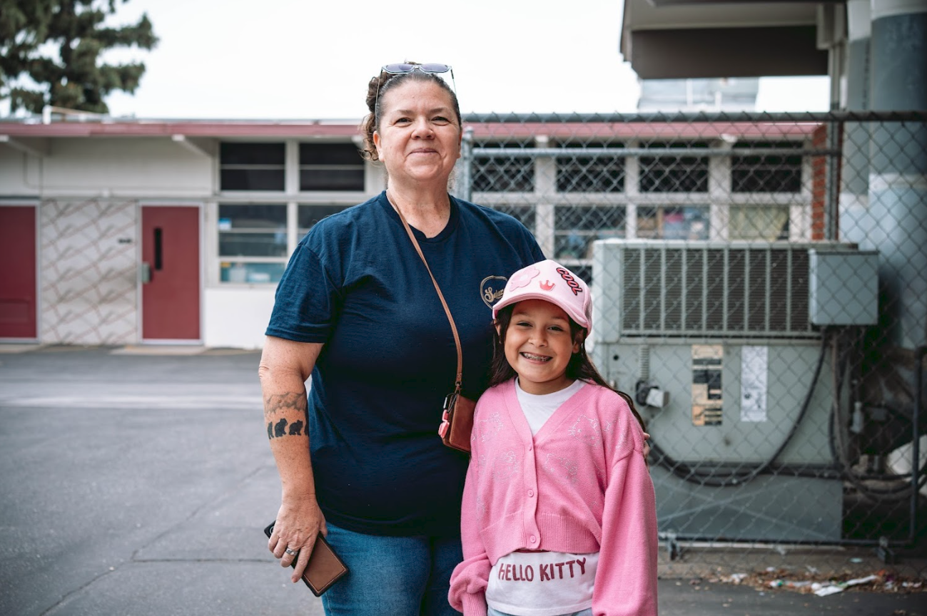 A woman and a young girl standing together on an outdoor pavement, smiling at the camera. The woman is wearing a dark blue shirt and has sunglasses on her head, while the girl is dressed in a pink jacket and a pink Hello Kitty hat. Behind them, there