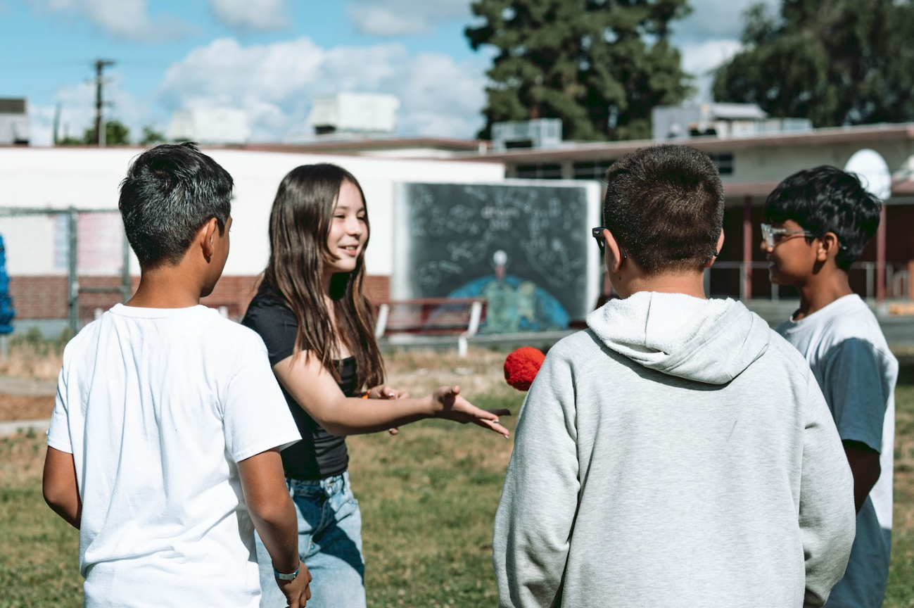 A group of four children standing outdoors on a sunny day, talking and playing with a red ball, with a school building and a chalkboard in the background.