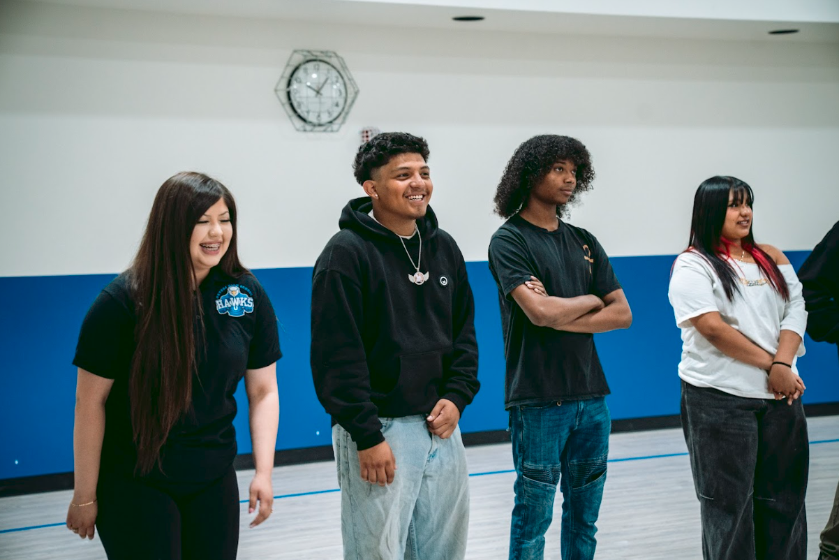 Four young people standing in a gymnasium, smiling and looking in different directions, with a clock on the wall behind them.