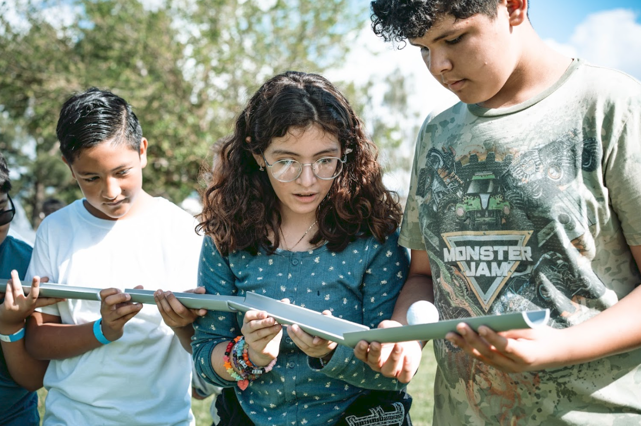 Group of children outdoors, looking at documents or worksheets, one boy is holding a golf ball.