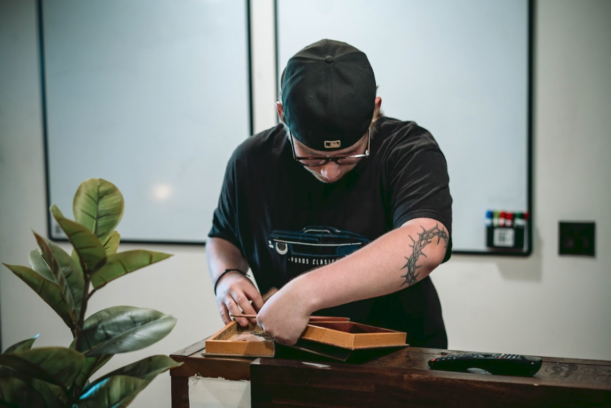 A man wearing a black baseball cap and glasses shows a tattoo on his arm as he puts items into a wooden box on a table in a room with a whiteboard in the background.