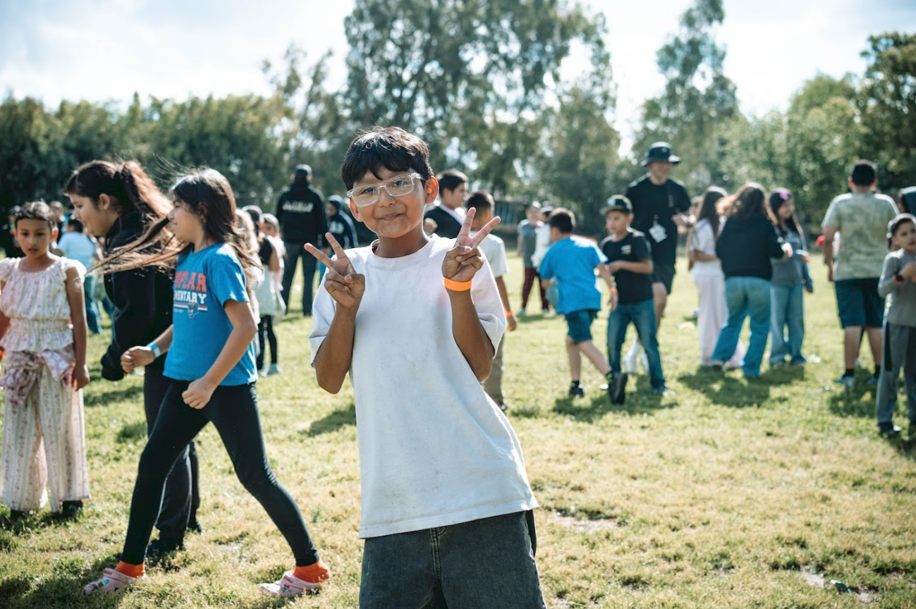 A young boy with glasses and a white shirt making peace signs with both hands at an outdoor event with many children and some adults on a grassy field.