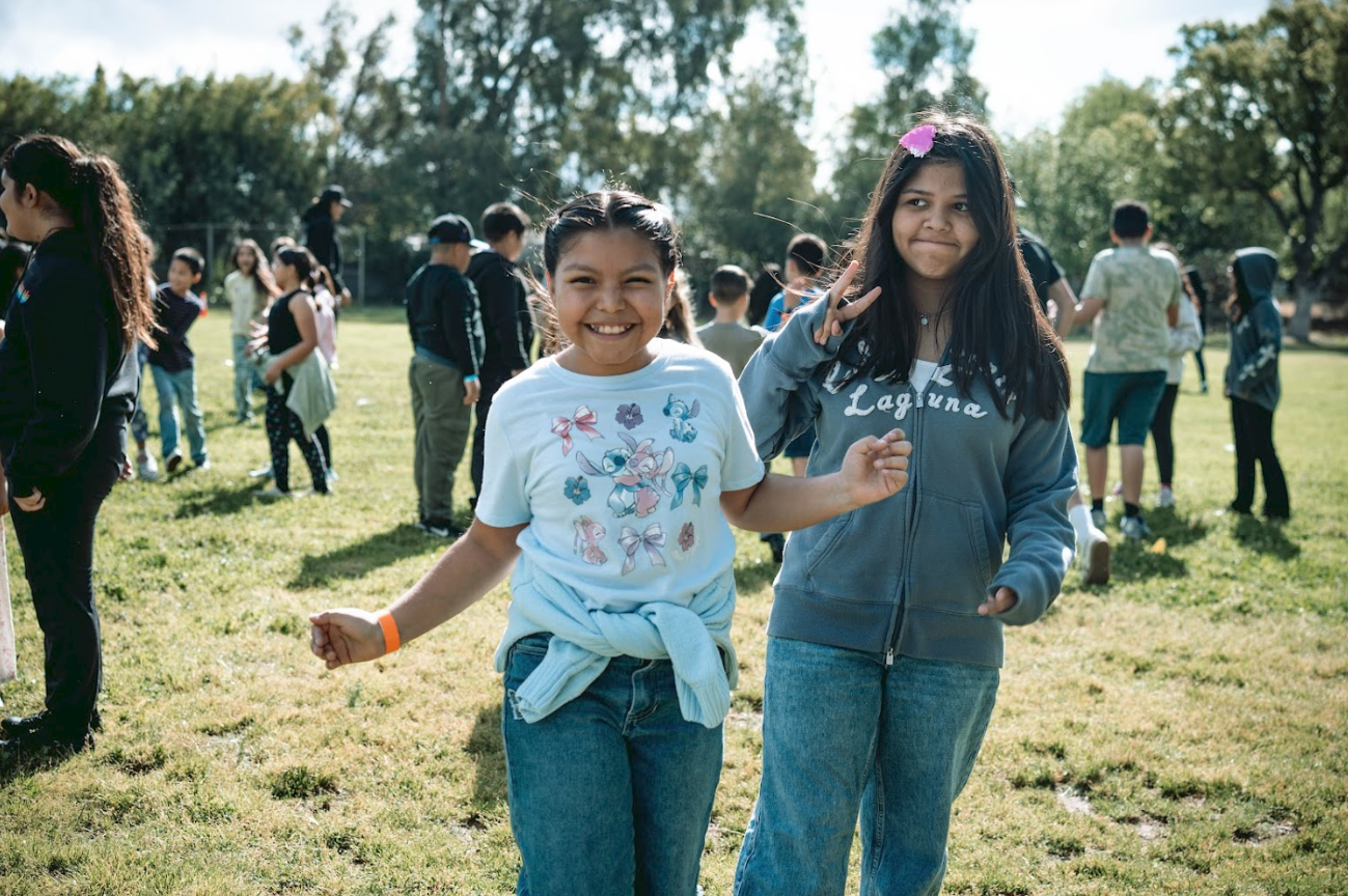 Two smiling girls, one with a bow shirt and the other with a hoodie, standing on a grassy field during an outdoor event, surrounded by other children and adults.