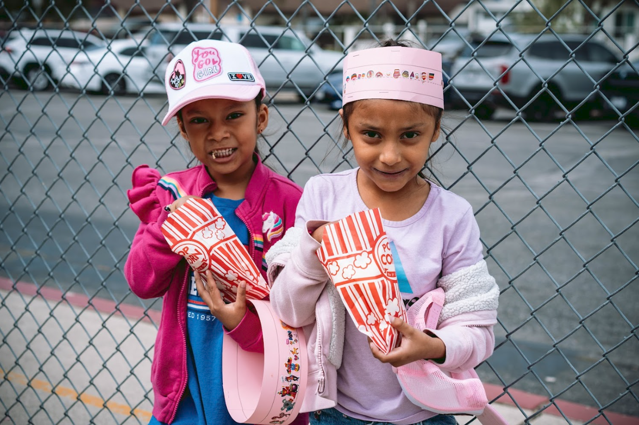 Two young girls standing behind a chain-link fence, each holding a box of popcorn and smiling at the camera.