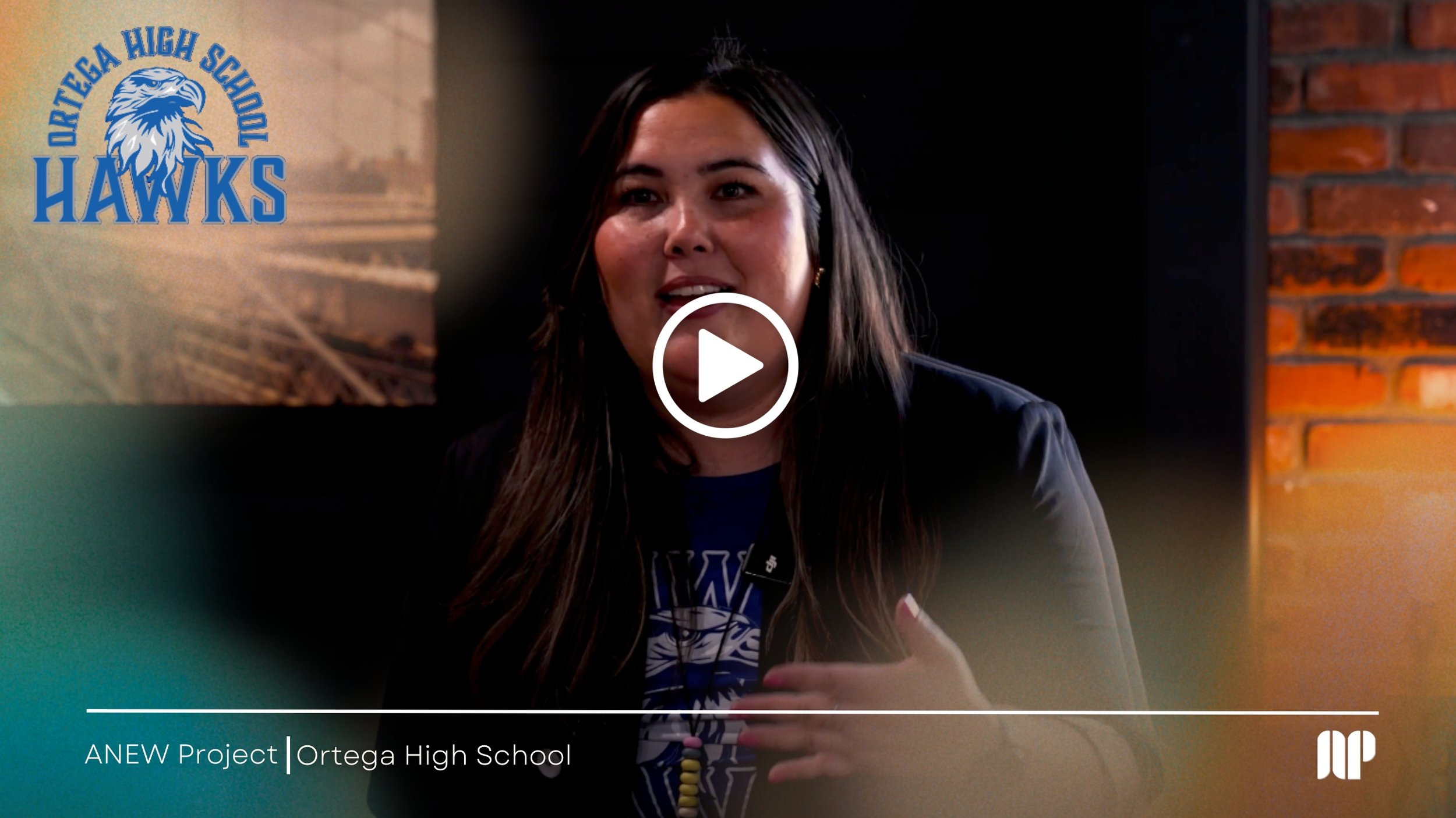 A woman with long dark hair speaking during a presentation in a room with brick walls, blue and green walls, and a logo on the wall that reads "Delta High School" with an eagle image. Text at the bottom mentions "ANEW Project Ortega High School."