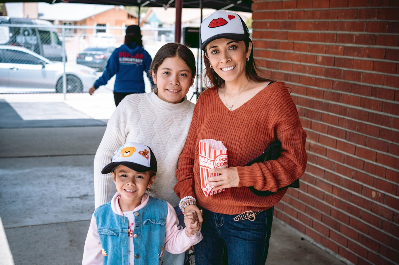 A woman and two young girls standing together outside a building, smiling. The woman holds a popcorn bag. Both girls are wearing baseball caps with popcorn and candy motifs. The woman wears a red sweater and the girls wear a white sweater and a pink 