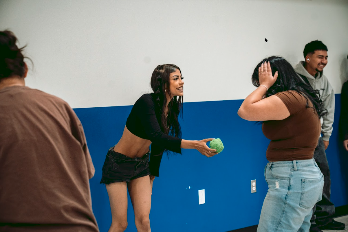 Two women exchanging a green stress ball, smiling and laughing, with others in the background in a room with a white and blue wall.