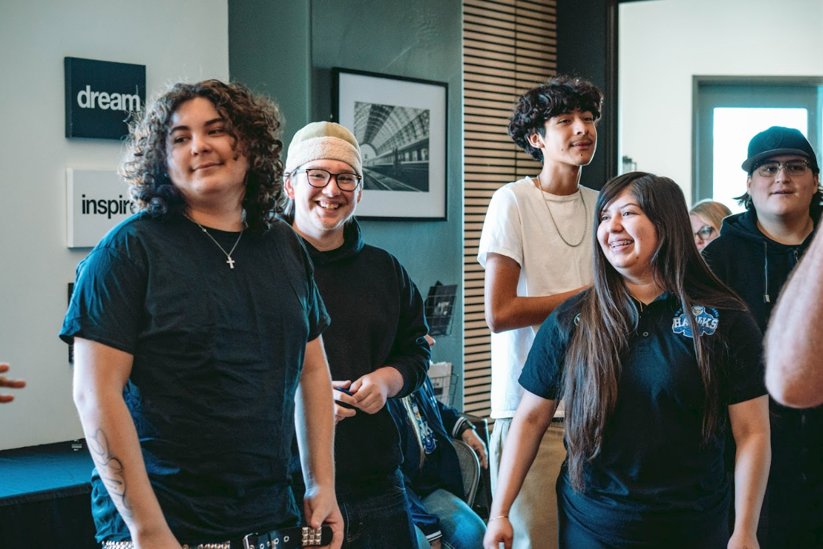 Group of five young people standing indoors, smiling and talking, with wall decorations and a door in the background.
