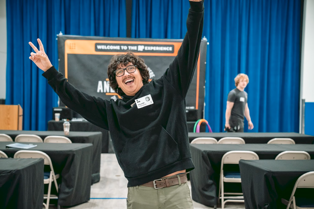 A person with curly hair and glasses smiling and raising their arms in a large conference room or auditorium, with a stage and a blue curtain backdrop behind them. There are empty tables with black tablecloths and chairs in the foreground, and anothe
