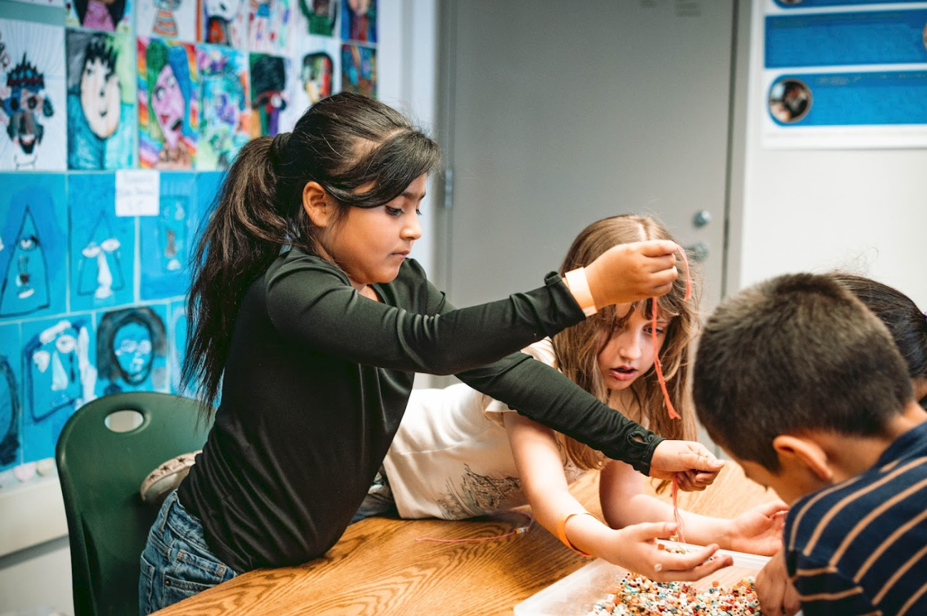 Children working together on a craft project at a table in a classroom, with colorful artwork displayed on the wall behind them.