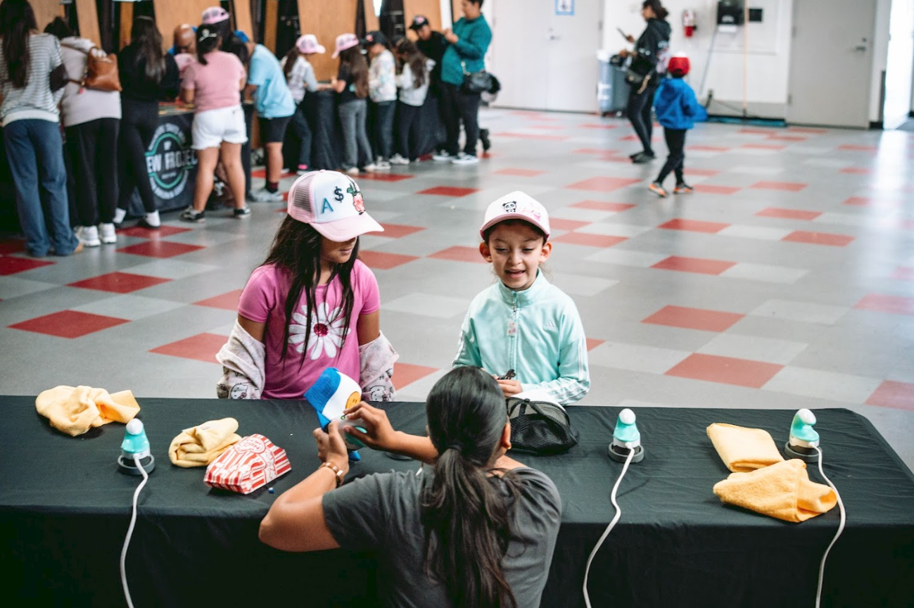 Children waiting in line and at a table where a woman is handing out small items at an indoor event or market.