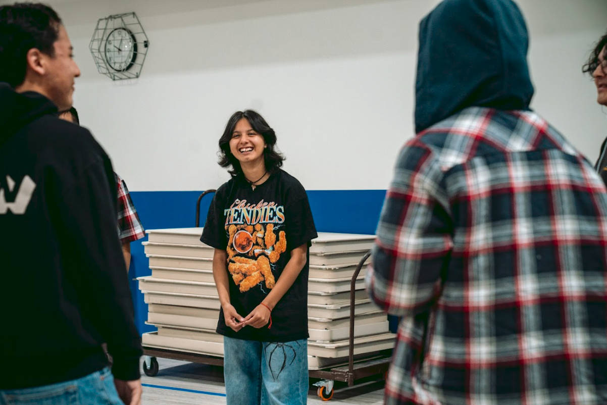 Group of young people talking and smiling in a gymnasium or community center, with tables stacked on carts in the background.
