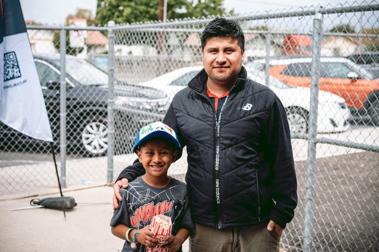 A man and a boy smiling at the camera in an outdoor parking lot. The boy is holding a popcorn bucket and wearing a colorful hat, while the man has his arm around the boy. Behind them, there is a chain-link fence and parked cars.